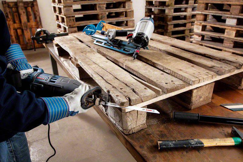 A person is sawing a wooden pallet with an electric saw. Stacked wooden pallets are visible in the background. Tools are lying on the table.