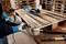 A person is sawing a wooden pallet with an electric saw. Stacked wooden pallets are visible in the background. Tools are lying on the table.