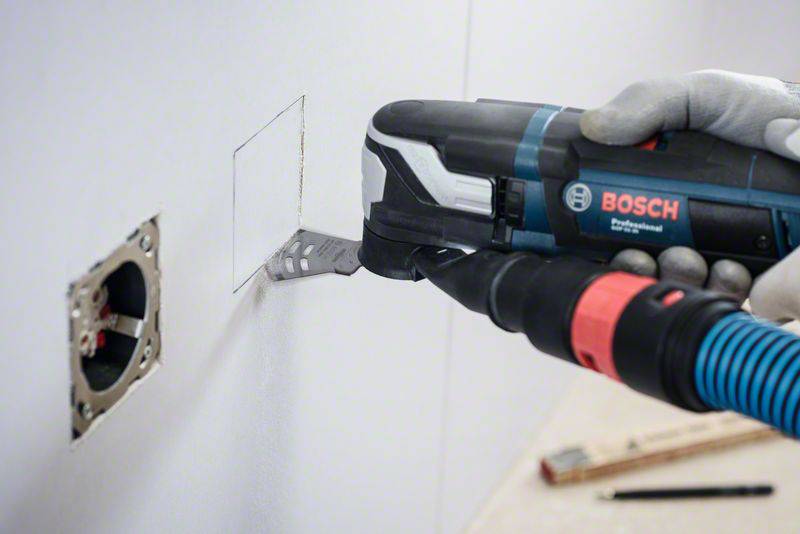 A person is precisely cutting a square into a white wall using an electric tool. A plug socket is visible nearby.