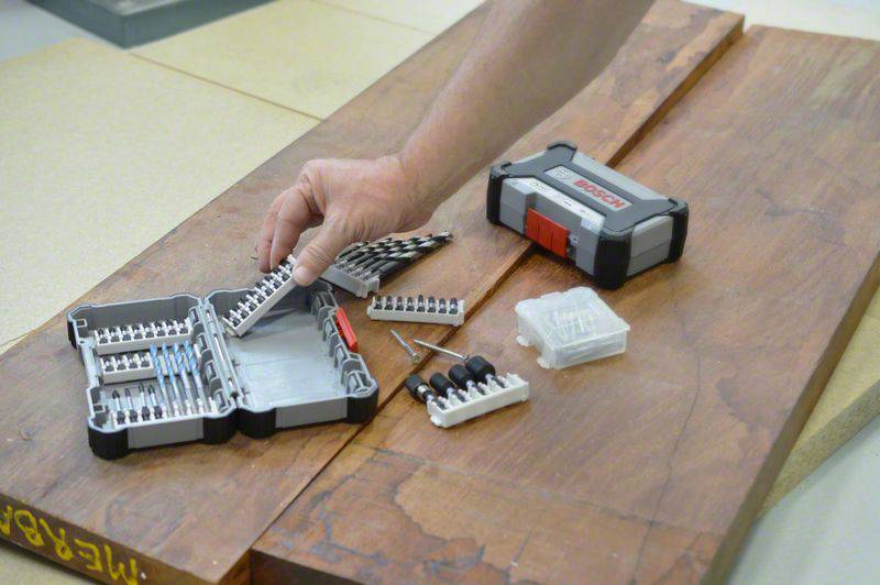 A hand reaches for a bit set on a wooden table. Various screwdriver bits and a box are visible. Ideal for woodworking.