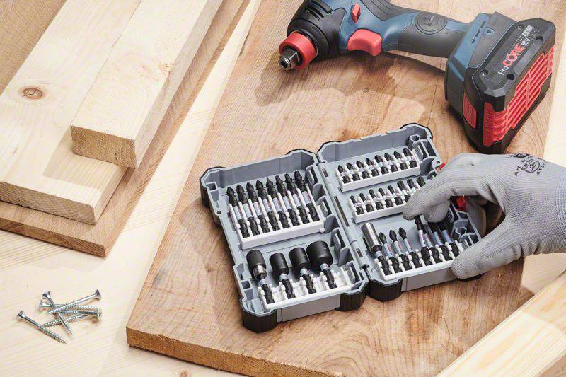 An open suitcase with drill bits on wooden boards, next to a cordless drill and loose-lying screws.