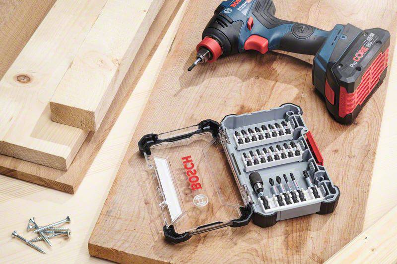 A cordless drill next to an open box of drill and screwdriver bits on wooden boards, with visible screws.
