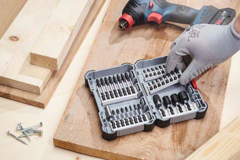 A person wearing gloves is sorting drill bits in an open toolbox on a wooden table. Planks and screws are lying nearby.