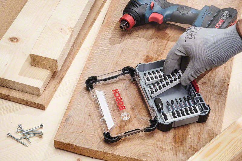 A person wearing a glove reaches for a drill bit in a Bosch tool case on a table with wooden boards and screws.