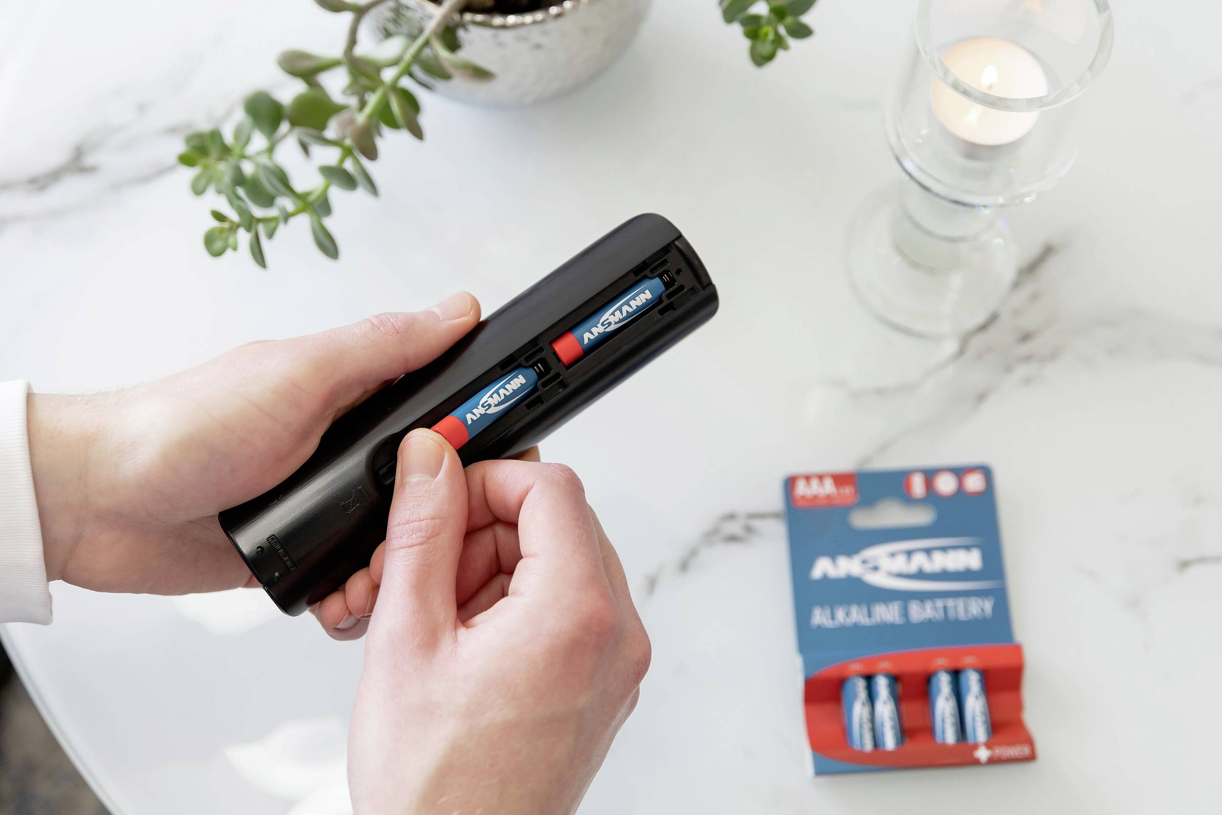 A person is inserting two alkaline batteries into a remote control. On the table are a pack of batteries, a plant, and a candle.
