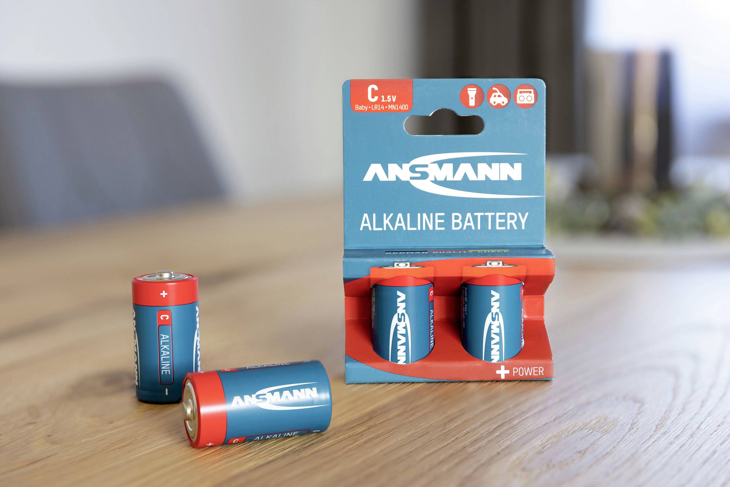 Three Ansmann C batteries on a table, two loose and one in its packaging. Room in the background slightly out of focus.