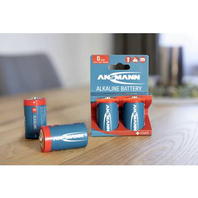 Four blue Ansmann alkaline batteries, Type D, on a table; two standing in their original packaging, two lying loose.