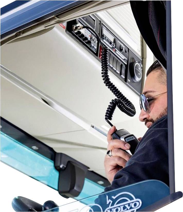 A man wearing sunglasses is speaking into a radio handset inside a vehicle cabin. Control panels and instrumentation are visible in the background.