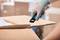 A person is cutting open a cardboard box with a black utility knife. The hand is wearing work gloves.