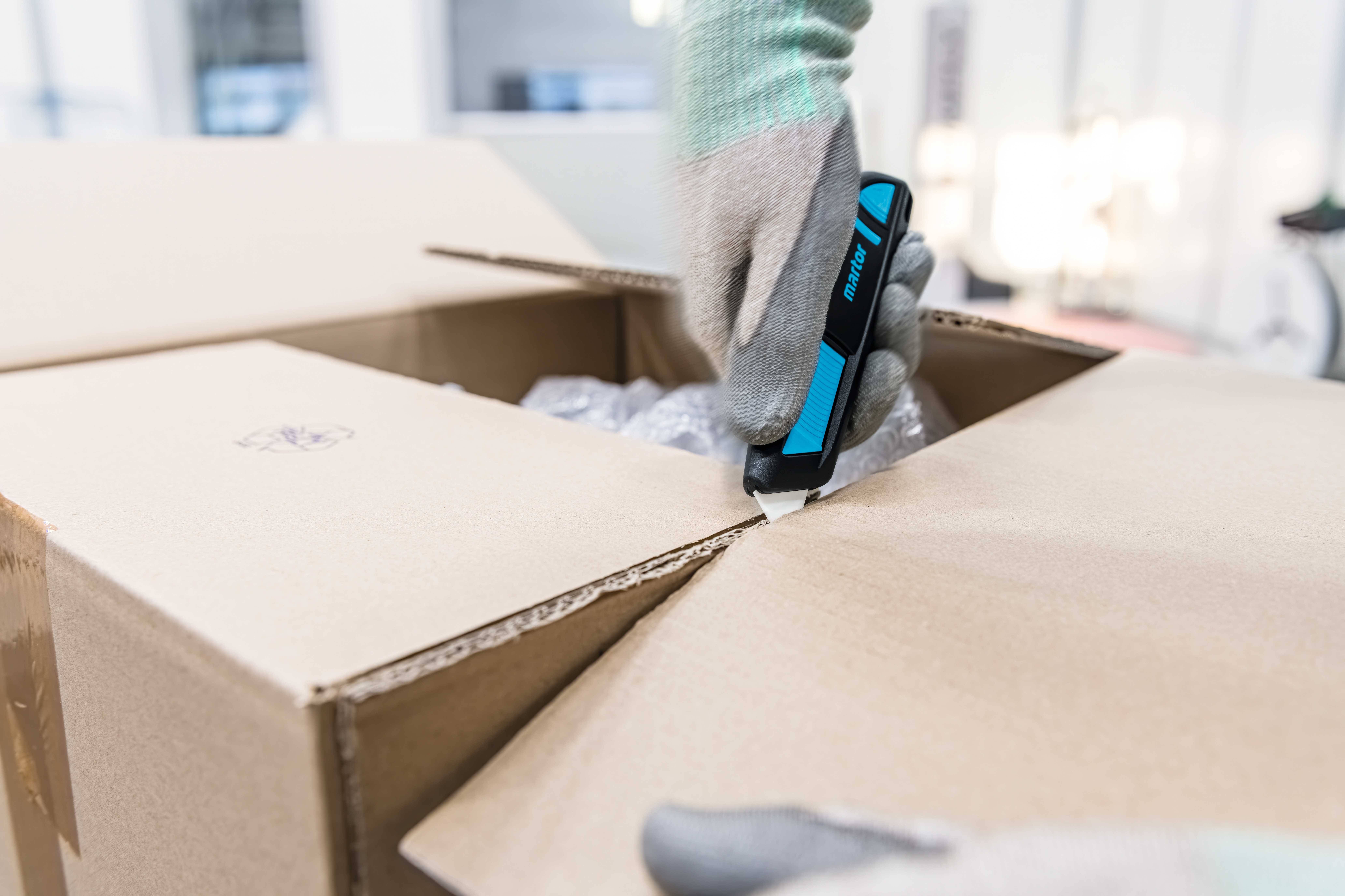 A hand wearing protective gloves opens a brown cardboard box with a blue utility knife. A bright room is visible in the background.
