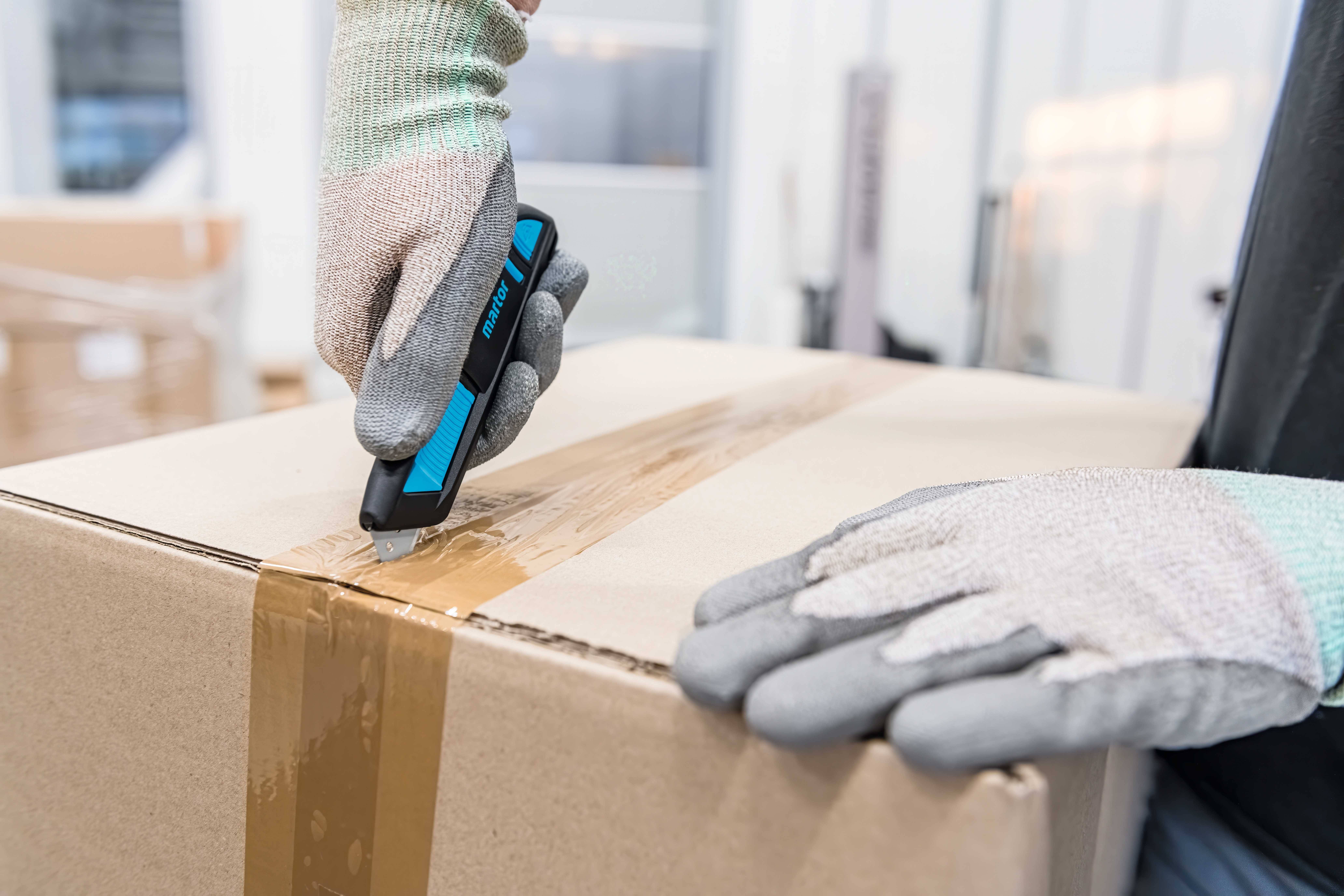 A person carefully opens a large parcel sealed with tape using a Stanley knife. They are wearing gloves.