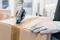 A person opens a cardboard box using a blue utility knife, protected by grey gloves. Blurry warehouse shelves are visible in the background.