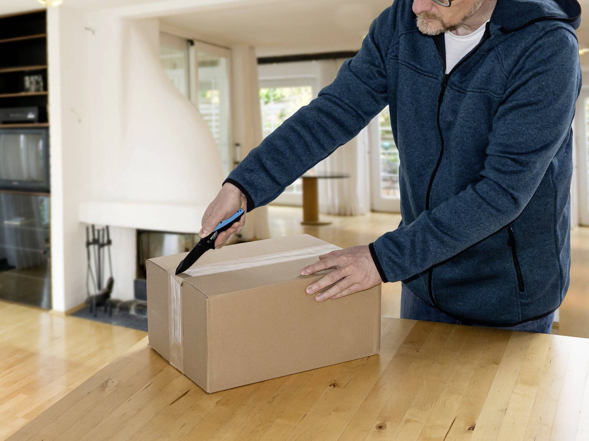 A person carefully opens a cardboard box with a knife on a wooden table in a living space.