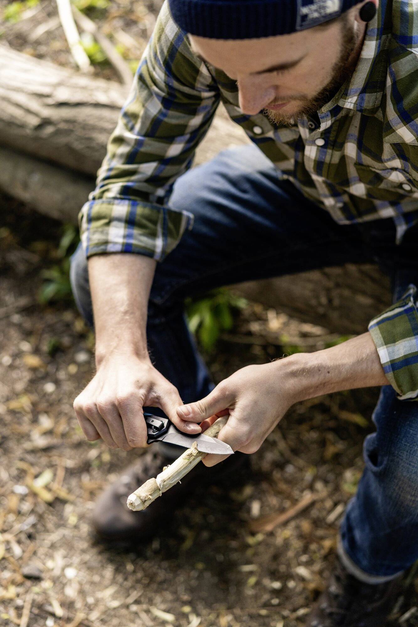A person is whittling with a knife on a stick outdoors. They are wearing a checked shirt and sitting near a tree trunk.