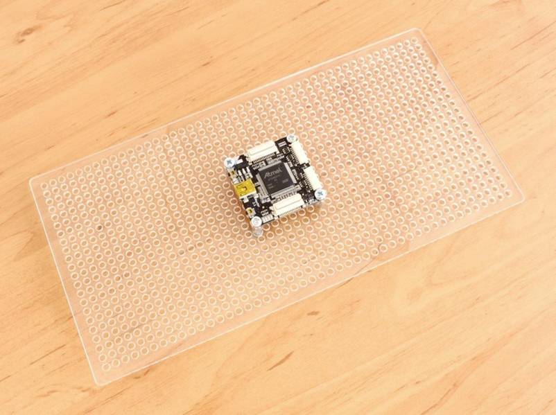 A microcontroller sits on a transparent breadboard, which is resting on a wooden surface.