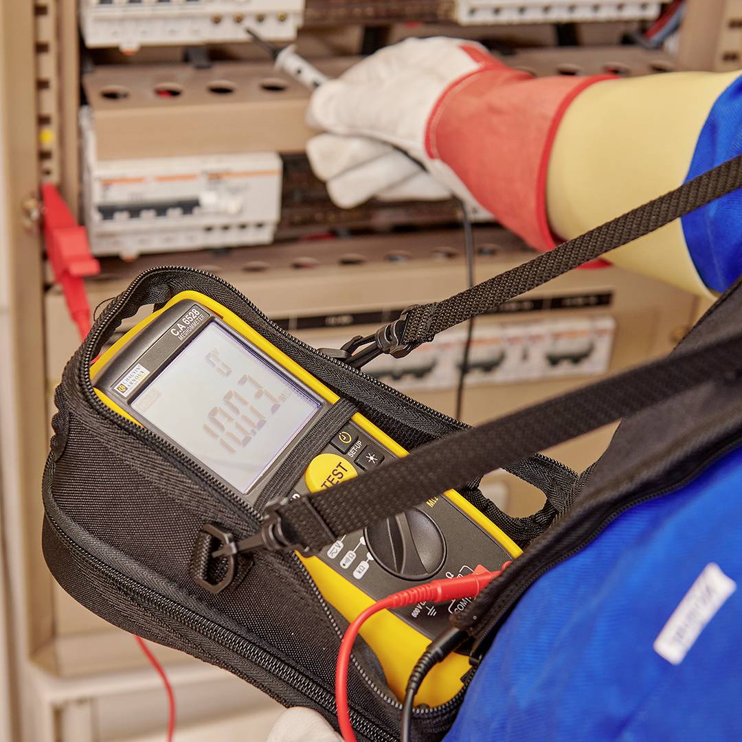 A person is measuring the voltage on an electrical control cabinet using a digital multimeter. The display shows '100.3'.<br><br>Note: In British English, decimal points are typically used instead of decimal commas, so I've changed '100,3' to '100.3'.