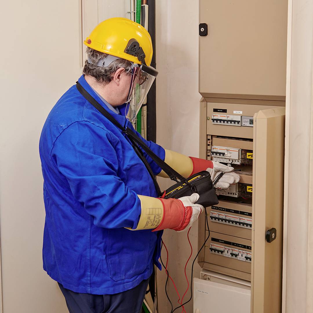 An electrician in blue workwear and a yellow hard hat is conducting tests on an open electrical cabinet using a multimeter.