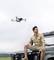 A man in beige clothing sits on a 4x4, gazing into the distance, while a drone hovers above him in the air.