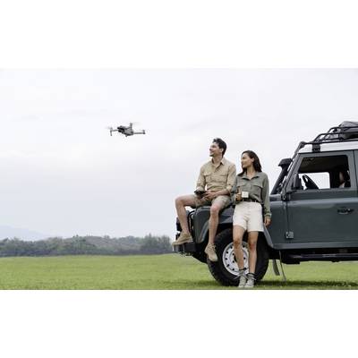A man and a woman are sitting in an off-road vehicle and controlling a drone over a vast, green field.
