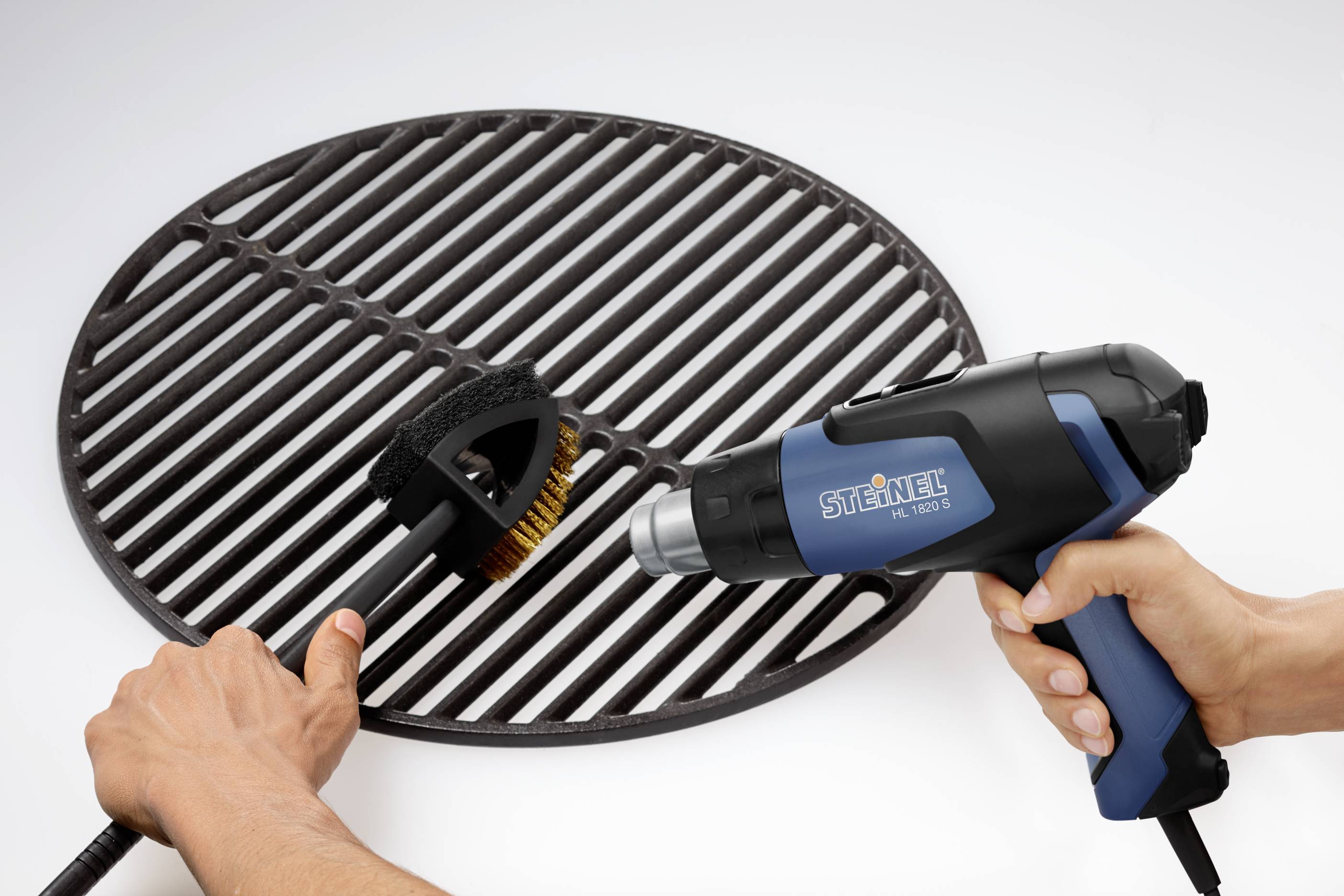 A person is cleaning a barbecue grill with a brush and a blue hot air blower.