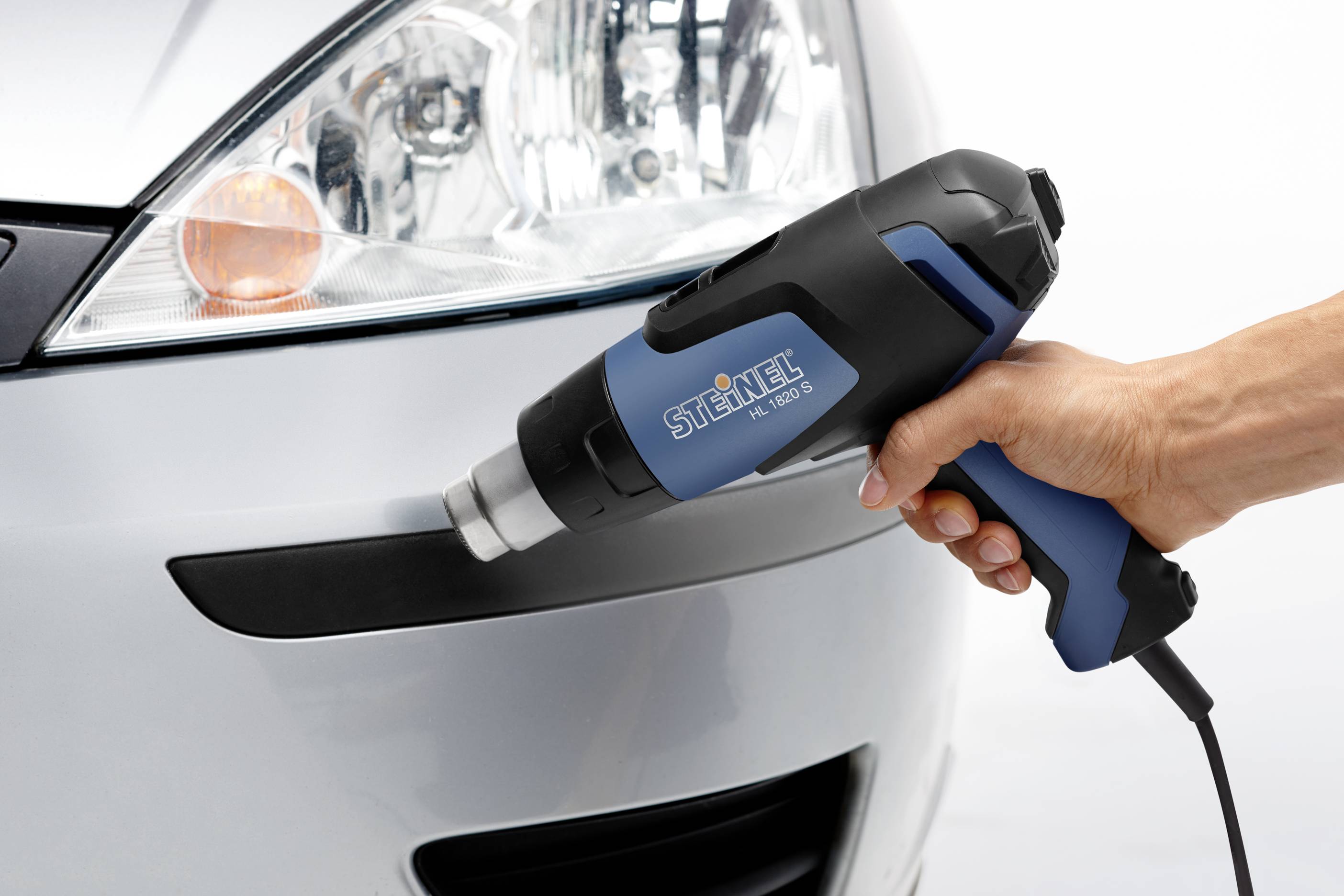 A hand holds a hot air blower near a car to shape a plastic bumper.