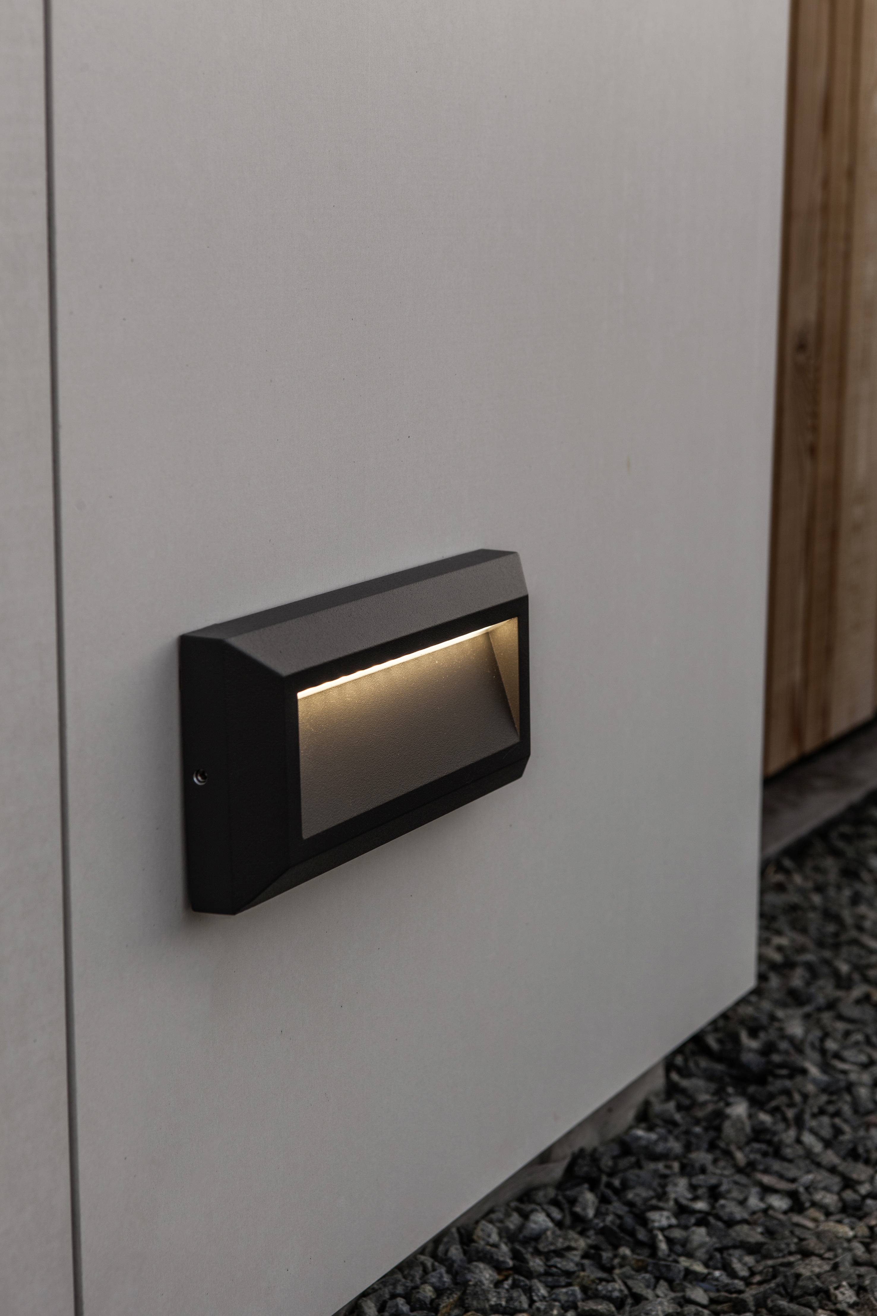 A black, rectangular exterior light is mounted on a light-coloured house wall, casting a dim light on the gravel path below.