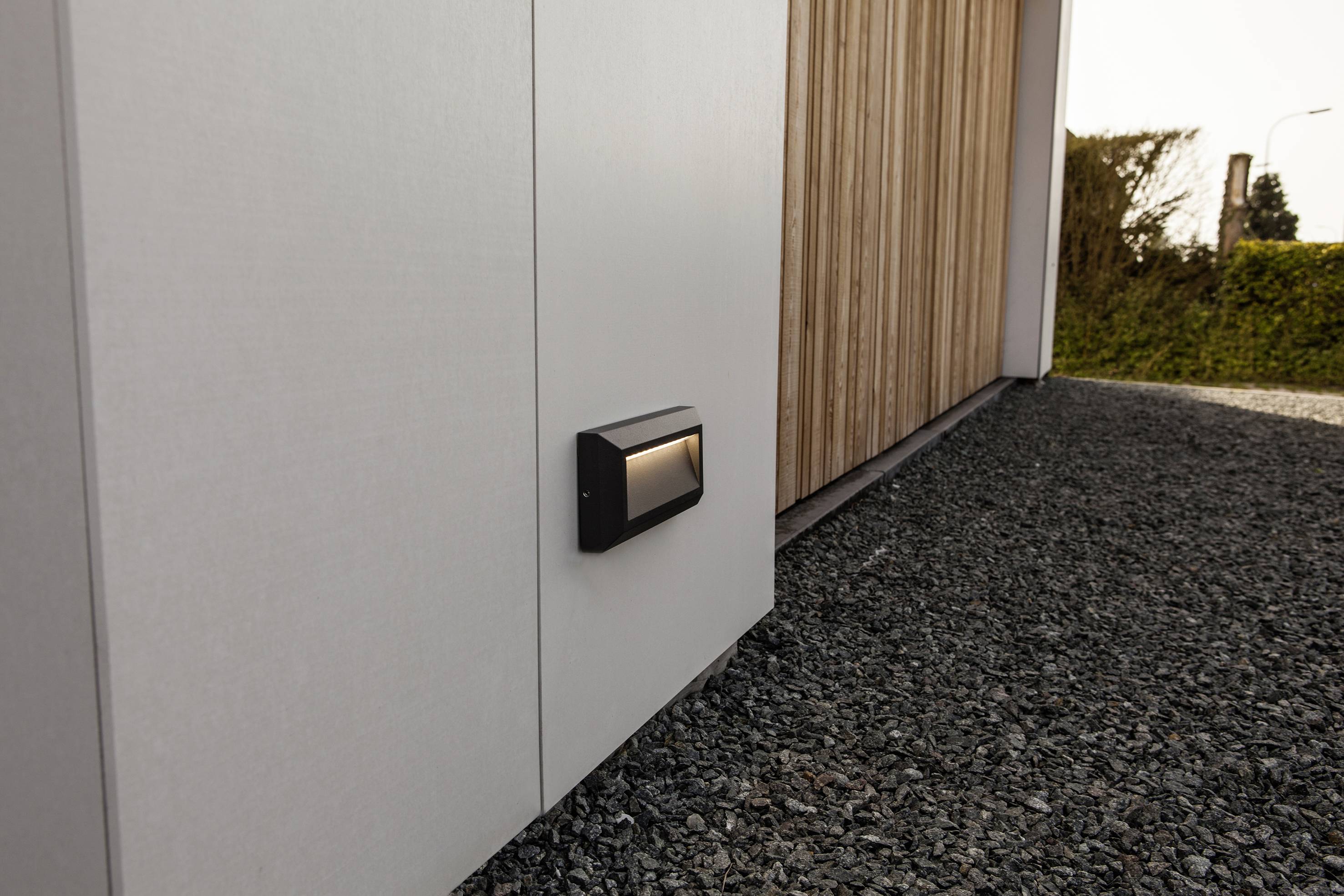 A grey building with wooden details and a modern wall light in a gravel-covered environment.