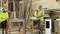 Two workers in yellow high-visibility vests are working in a woodworking workshop, surrounded by stacks of timber and tools on a workbench.