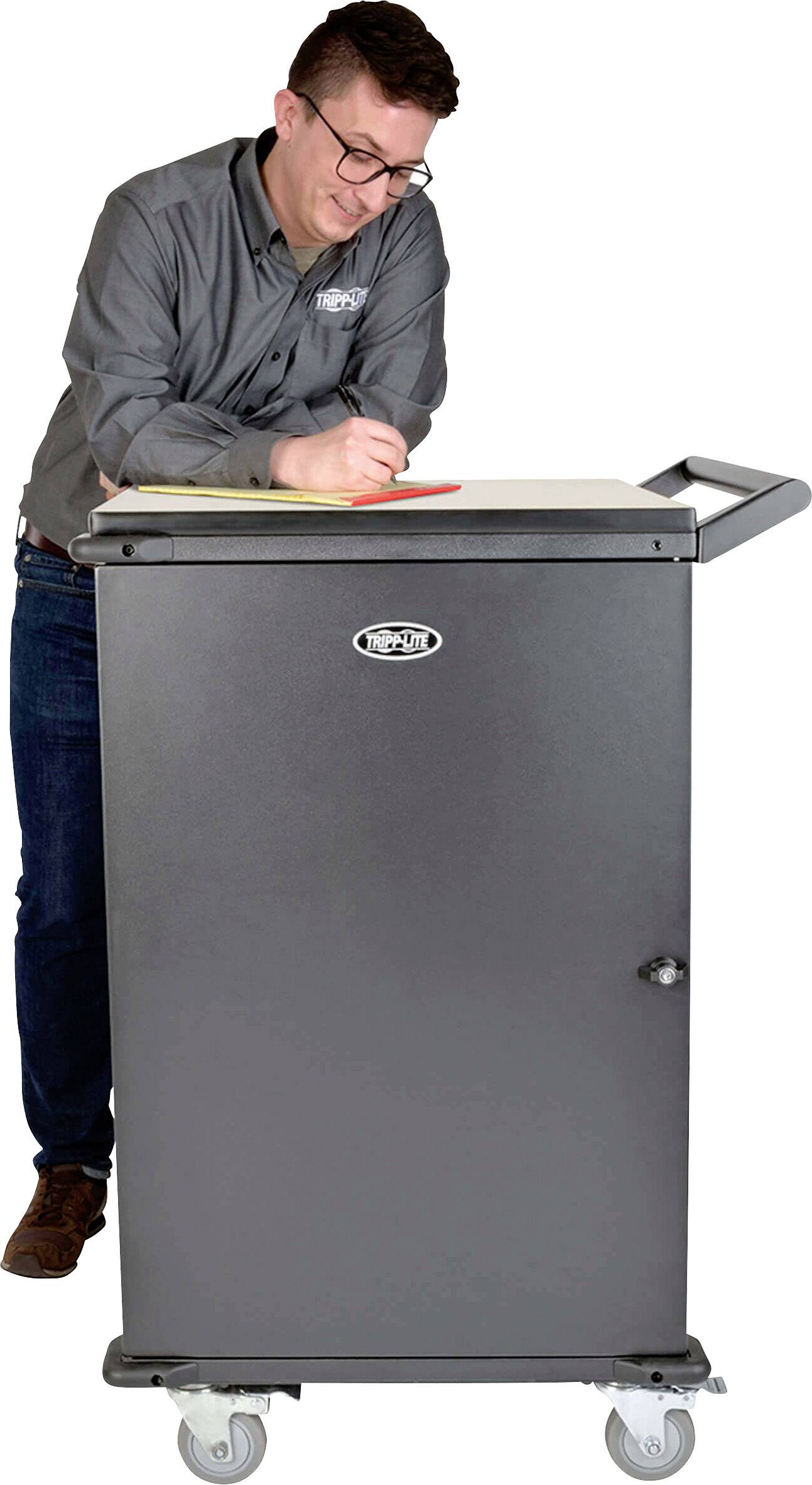 A man is standing next to a large, grey cabinet on wheels and writing in a notebook. The cabinet has a circular lock.