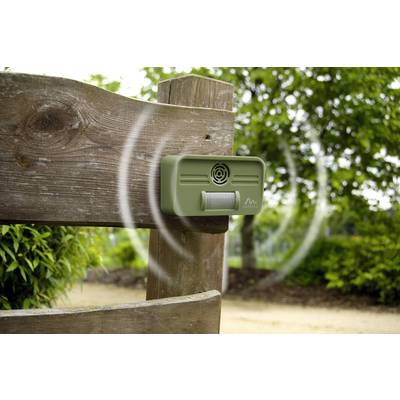 An ultrasonic animal deterrent is attached to a wooden fence in the garden, emitting sound waves. Trees and shrubs are visible in the background.
