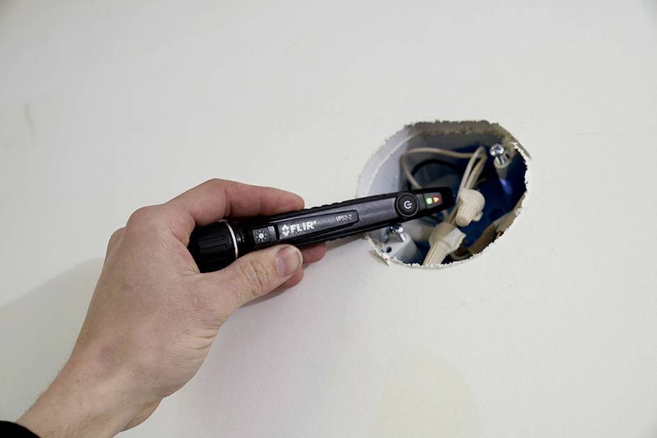 A person is holding an electrical voltage tester against an exposed cable in a wall opening to check electrical safety.