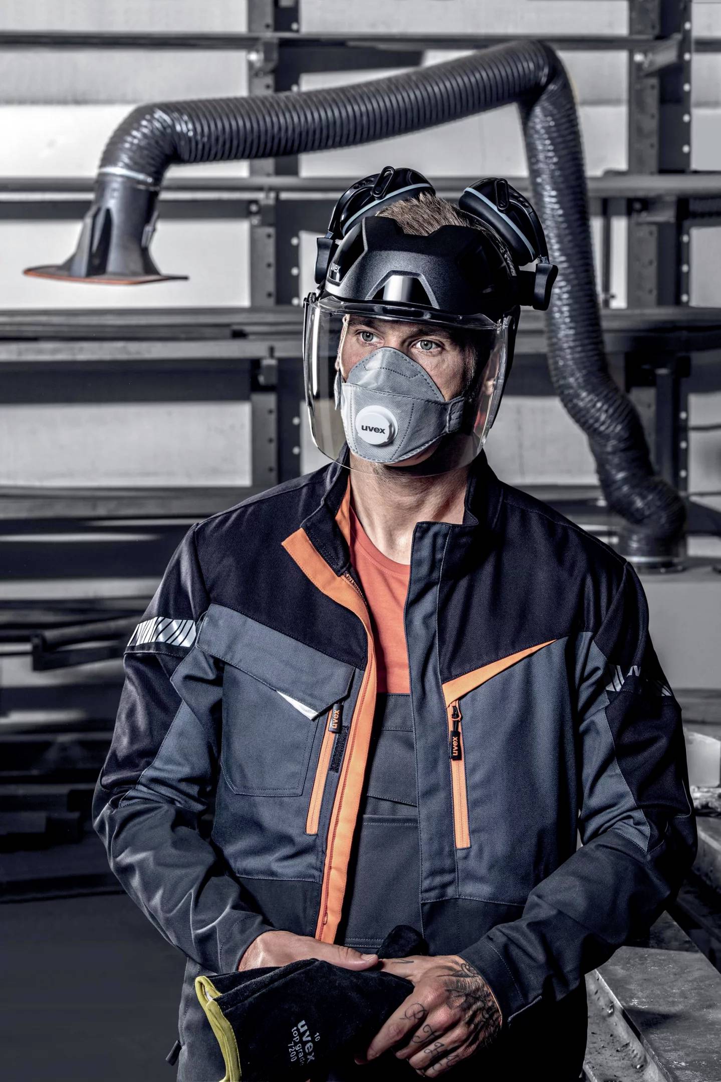 Worker wearing protective clothing and a respiratory mask stands in an industrial environment. Background shows ventilation ducts.