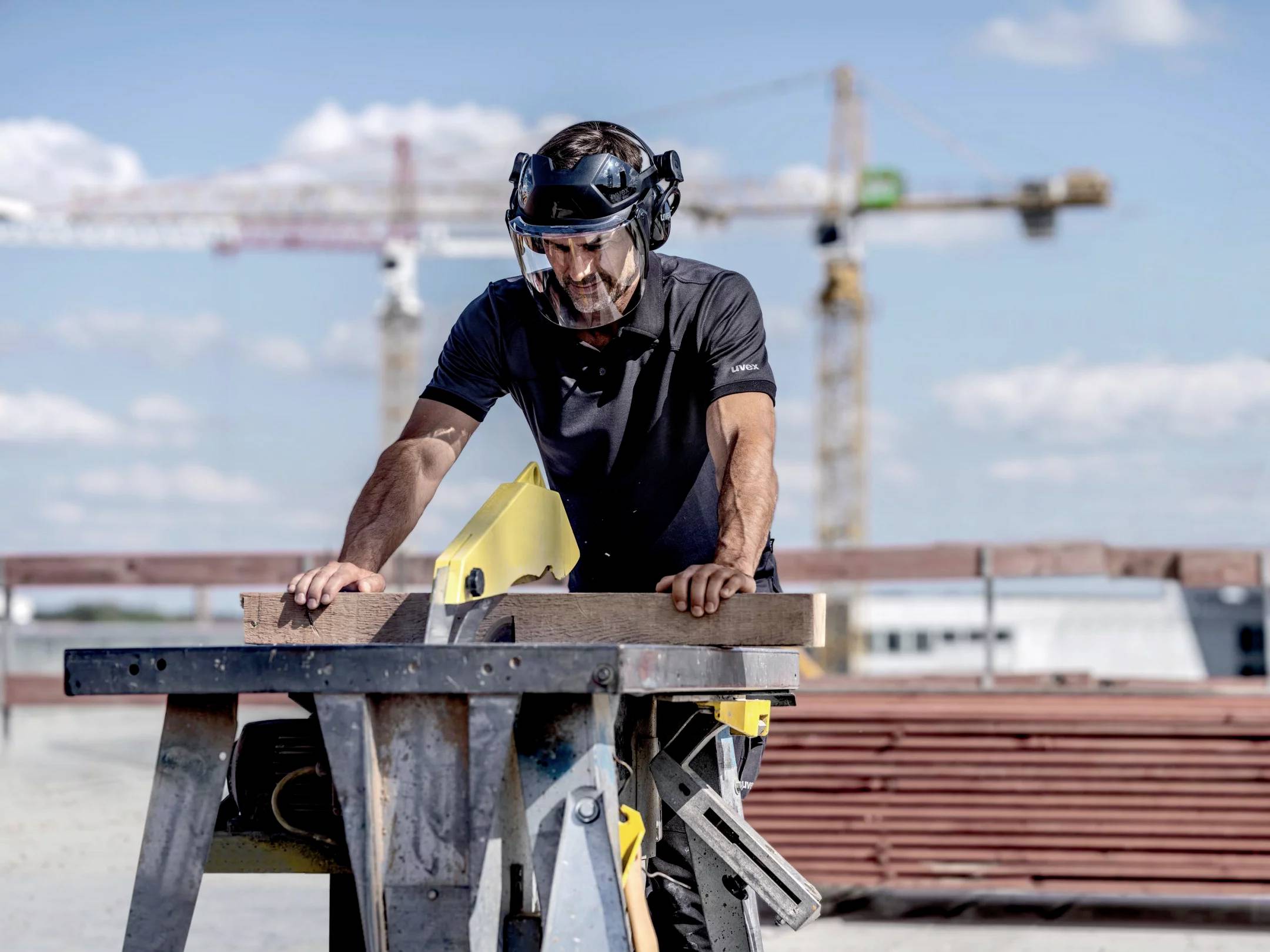 A man is working with a circular saw on an outdoor construction site. Construction cranes are visible in the background.