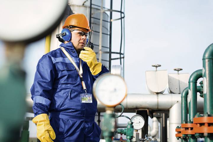A worker in safety clothing and a hard hat is checking an installation in an industrial facility using a two-way radio. He is wearing ear protection.