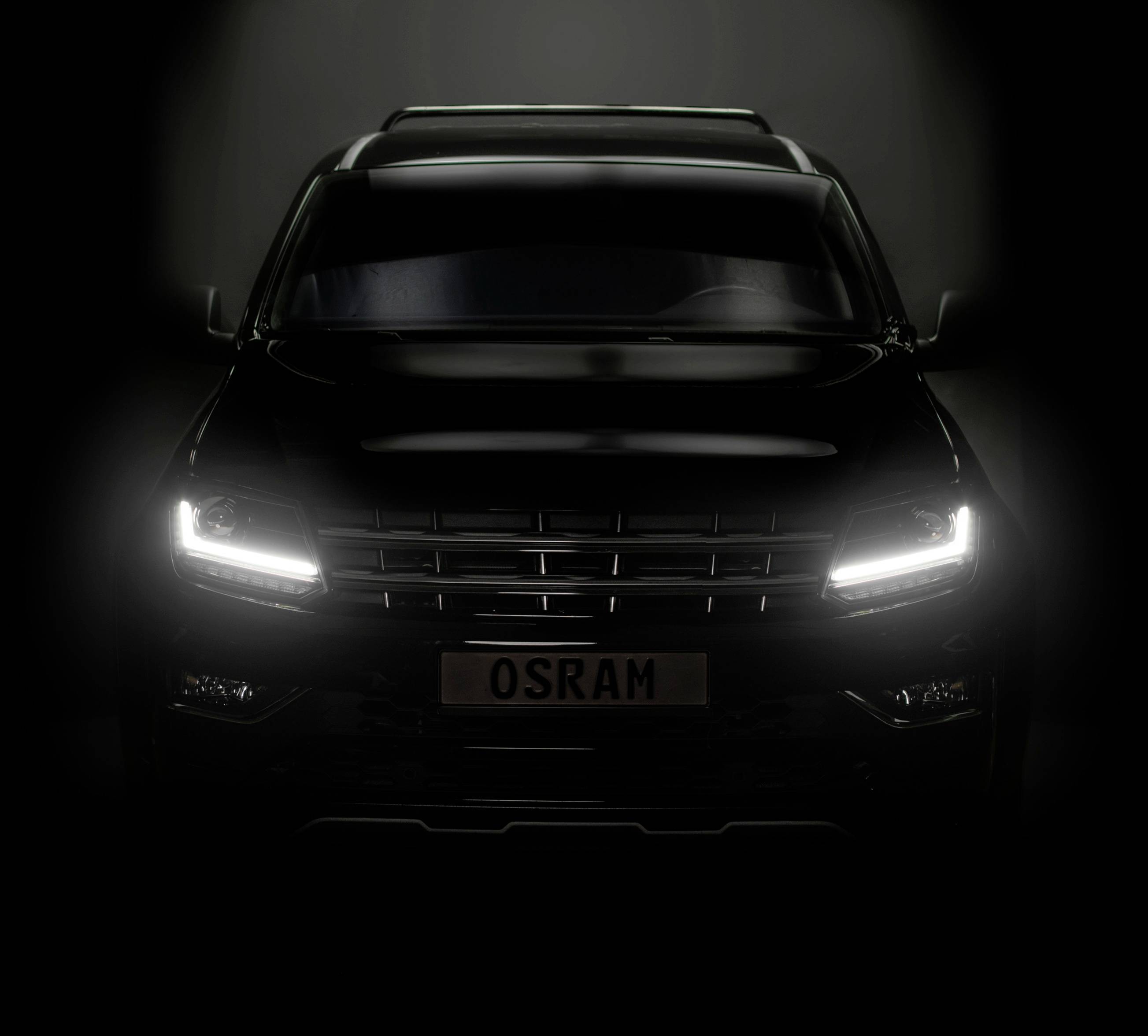 A black car with illuminated headlights drives at dusk. The number plate reads 'OSRAM'.