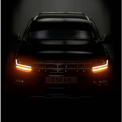 A black car with illuminated headlights, shown head-on in the darkness. The number plate reads 'OSRAM'.