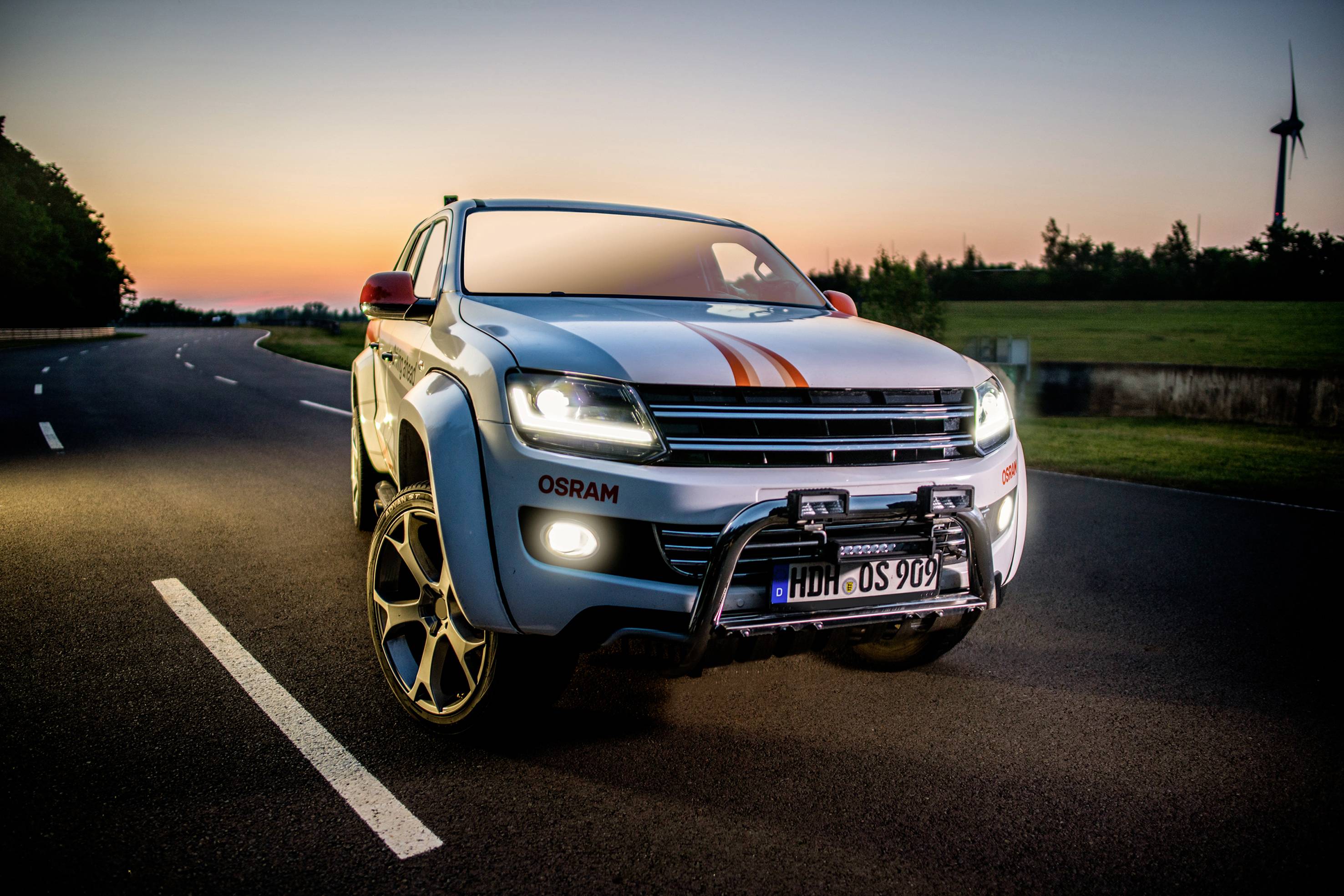A white car with racing stripes on a country road at sunset. A wind turbine is visible in the background.