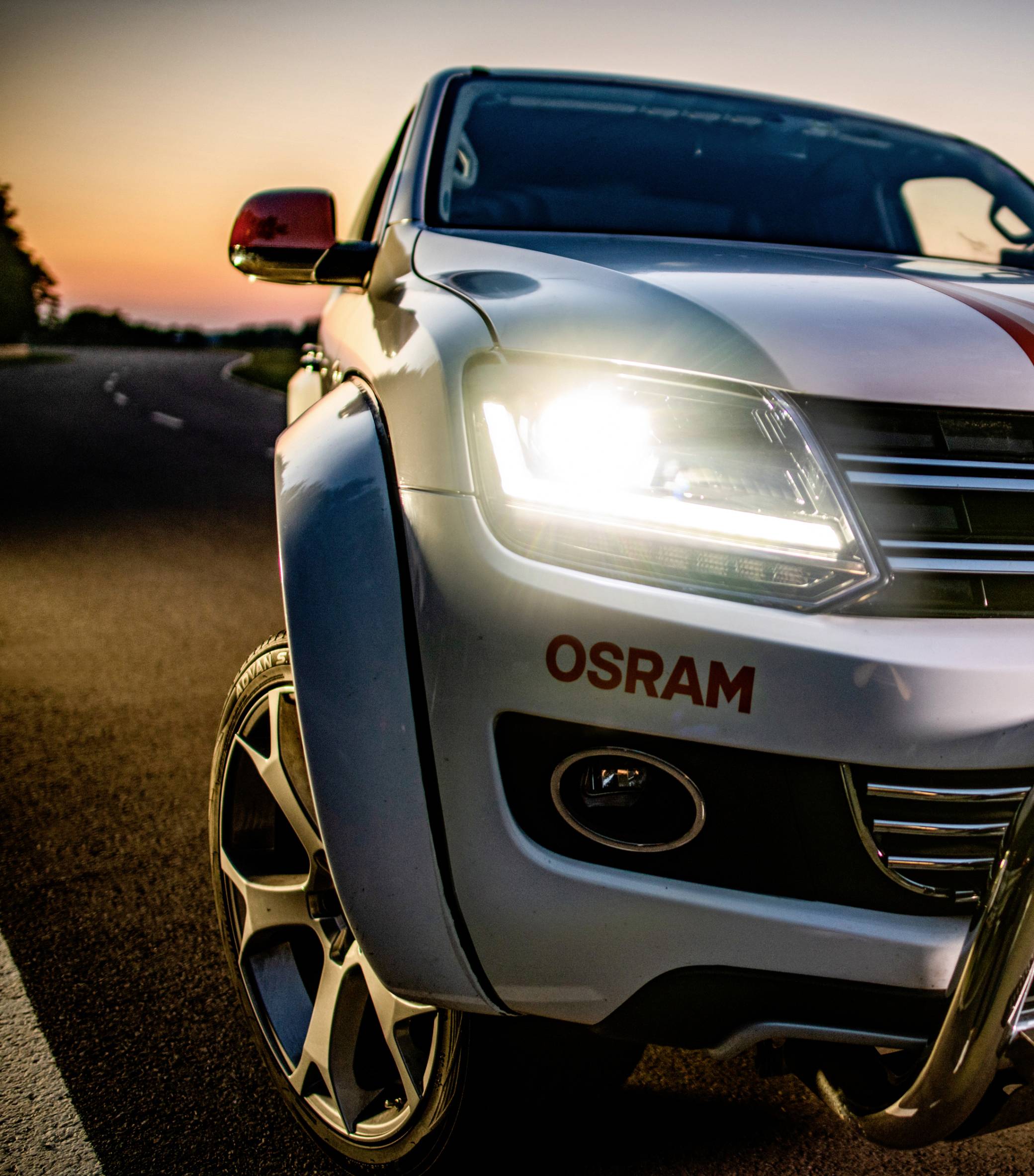 A car with its headlights on drives along a country road at sunset.
