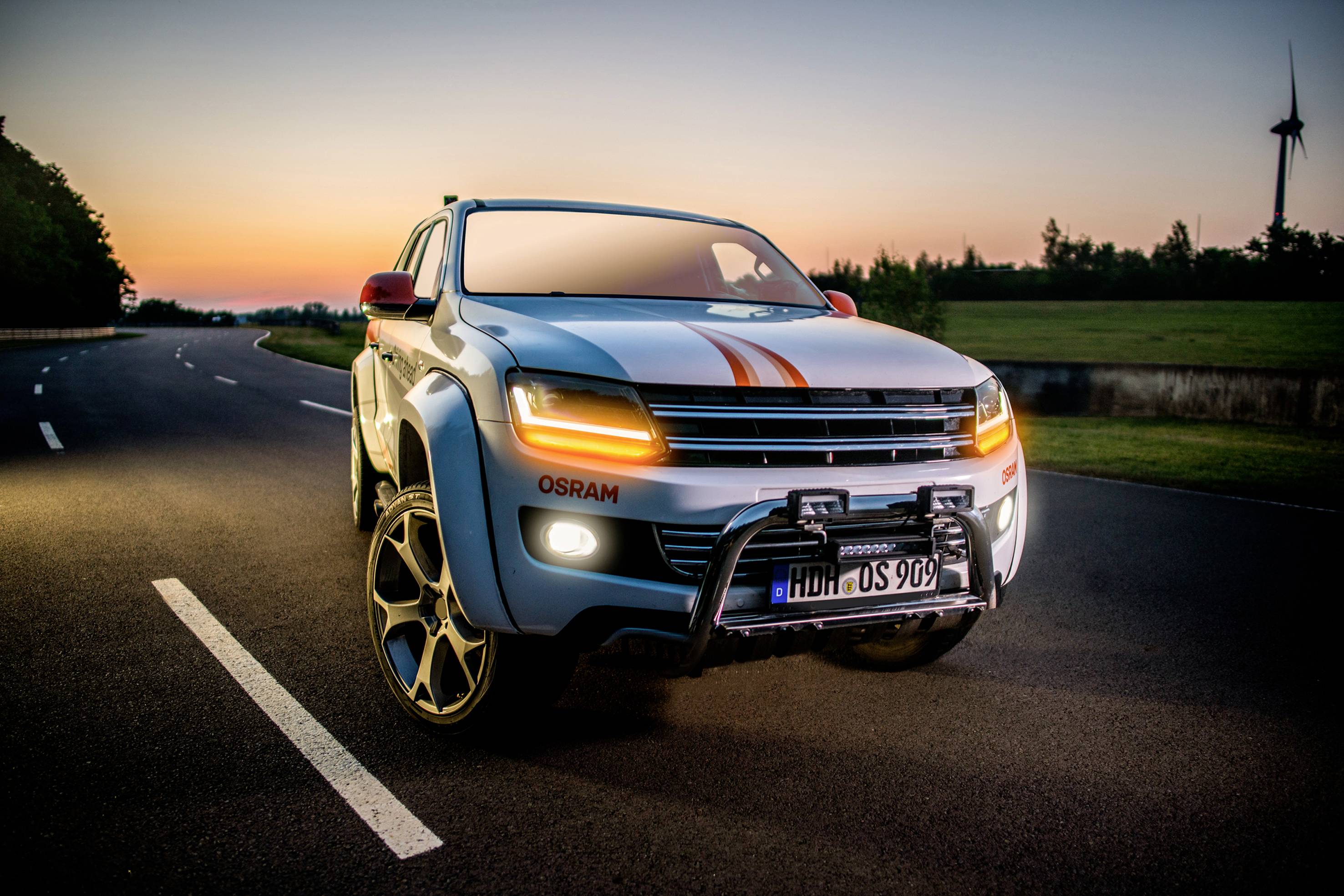 Off-road vehicle with illuminated headlights driving on a country road at sunrise. Wind turbine in the background.