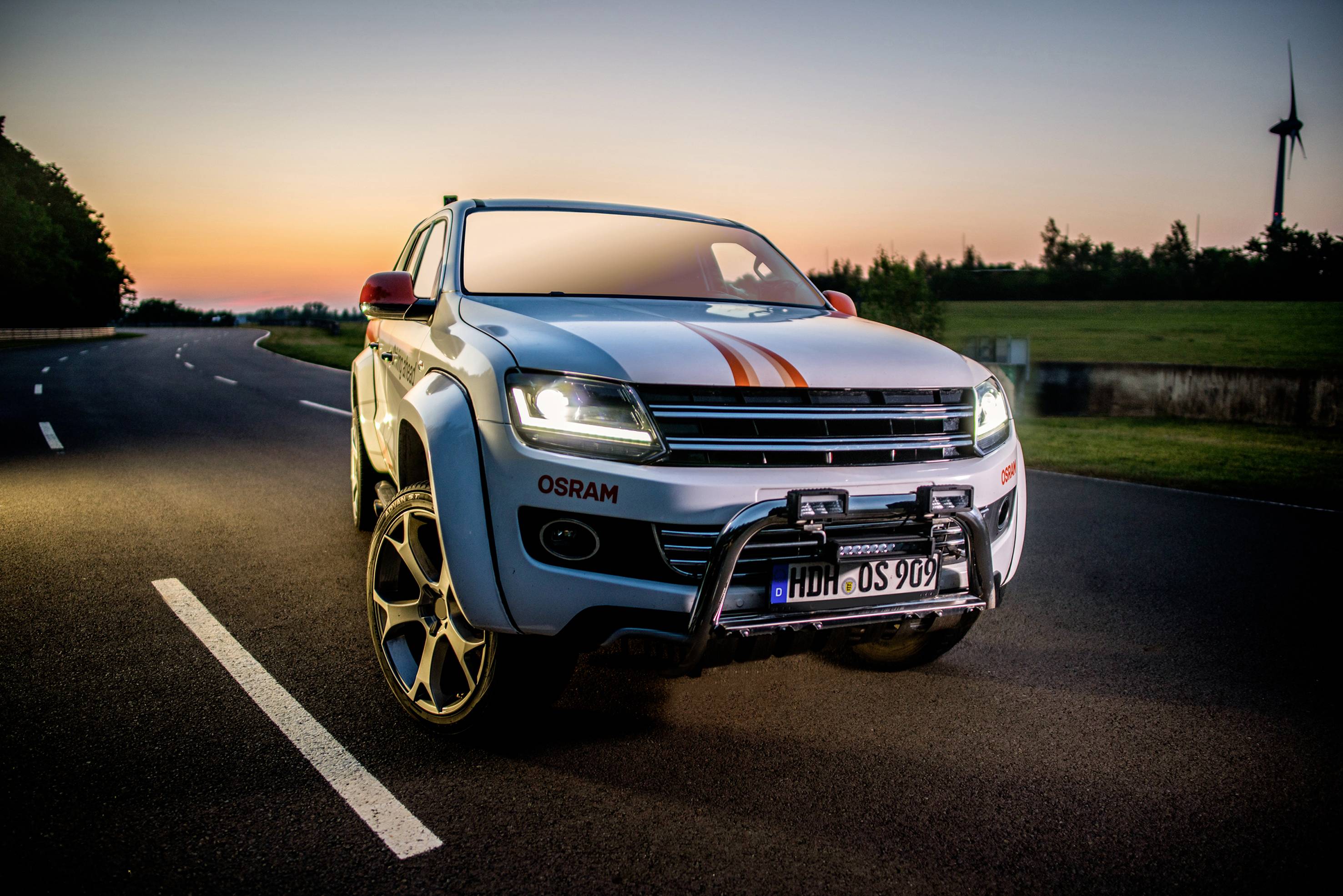 A white SUV with stickers and additional headlamps is parked on an empty country road at sunset, with a wind turbine in the background.