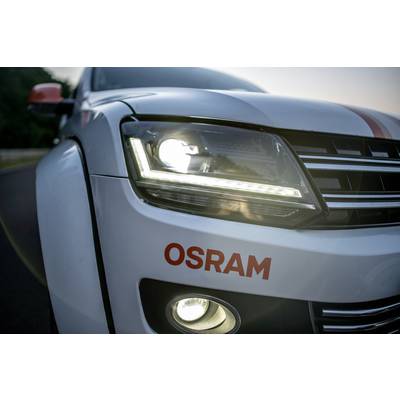 White car with OSRAM inscription, headlights and fog lights illuminated on a road.