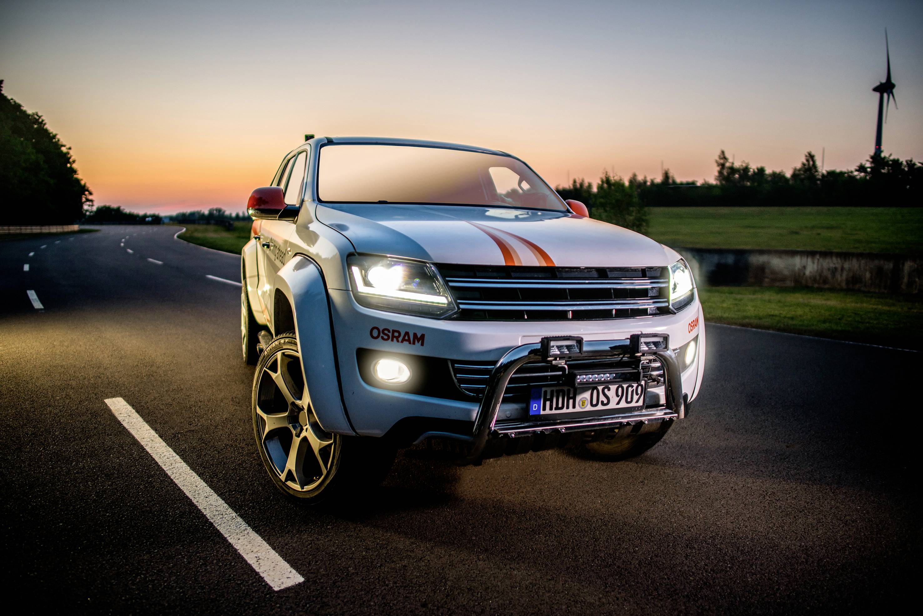 An off-road vehicle with racing stripes is parked on a road at sunset, with a wind turbine in the background.