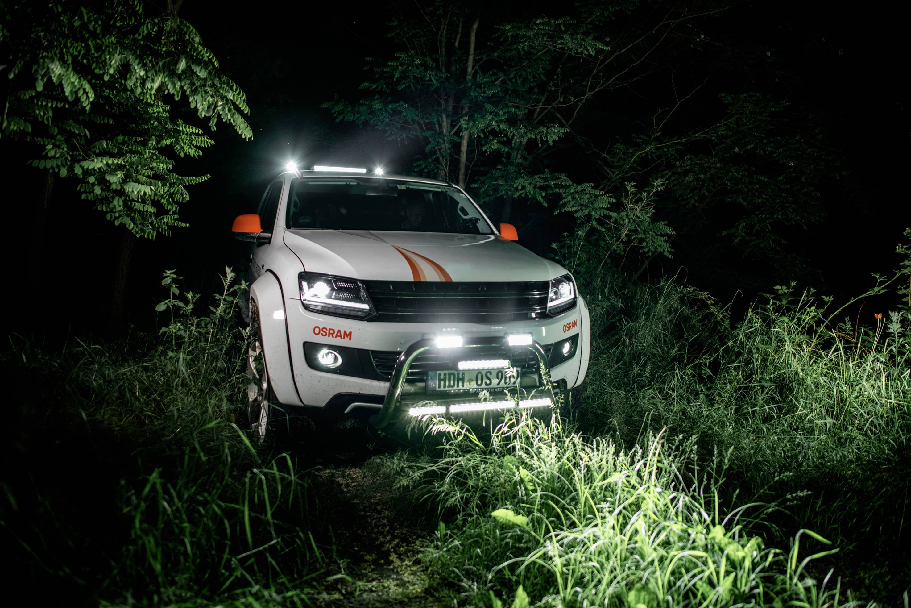 A white off-road vehicle with orange accents is parked at night on a tree-lined path, surrounded by trees, with its lights on.