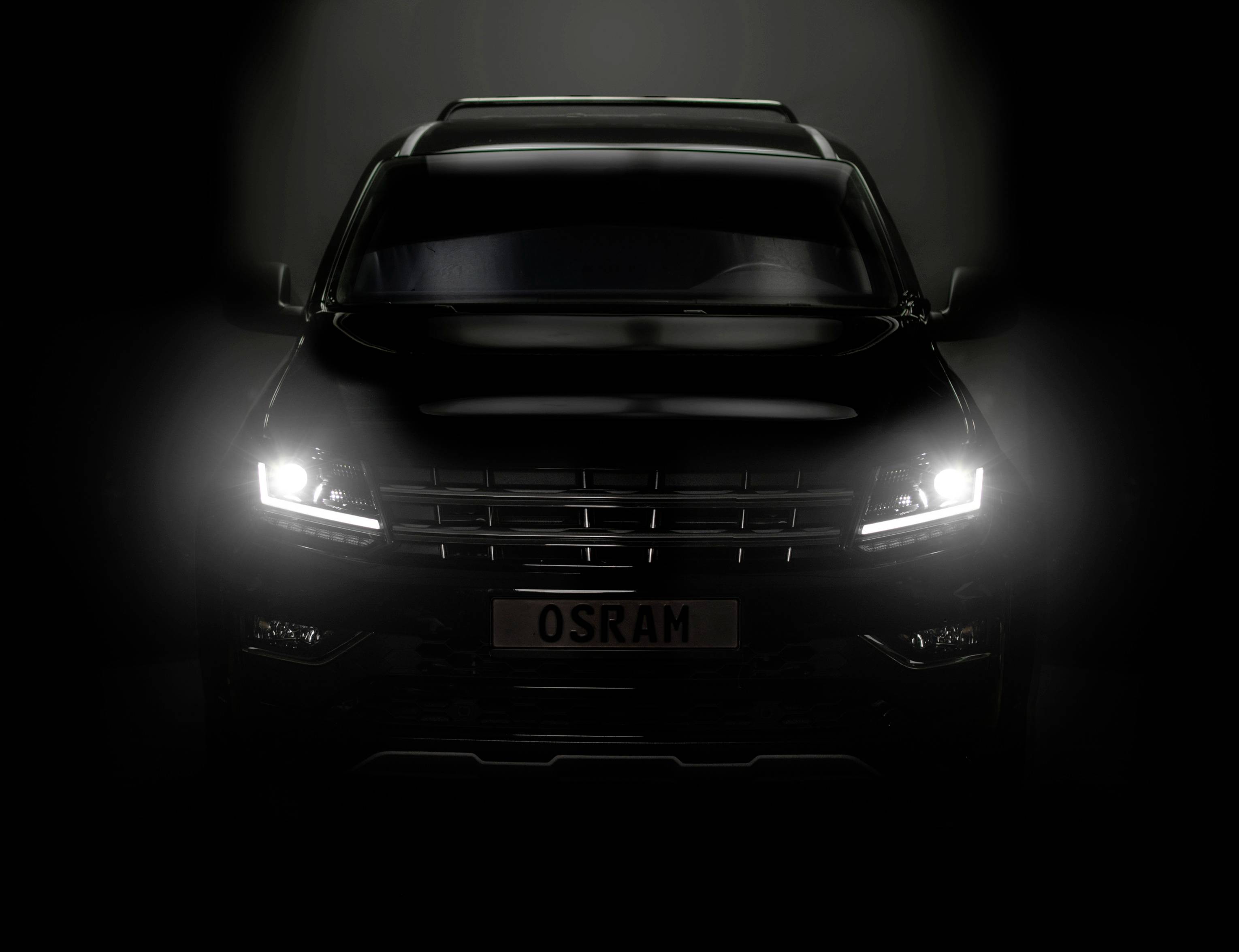 A black car with headlights switched on gleams in a dark environment. The number plate reads 'OSRAM'.