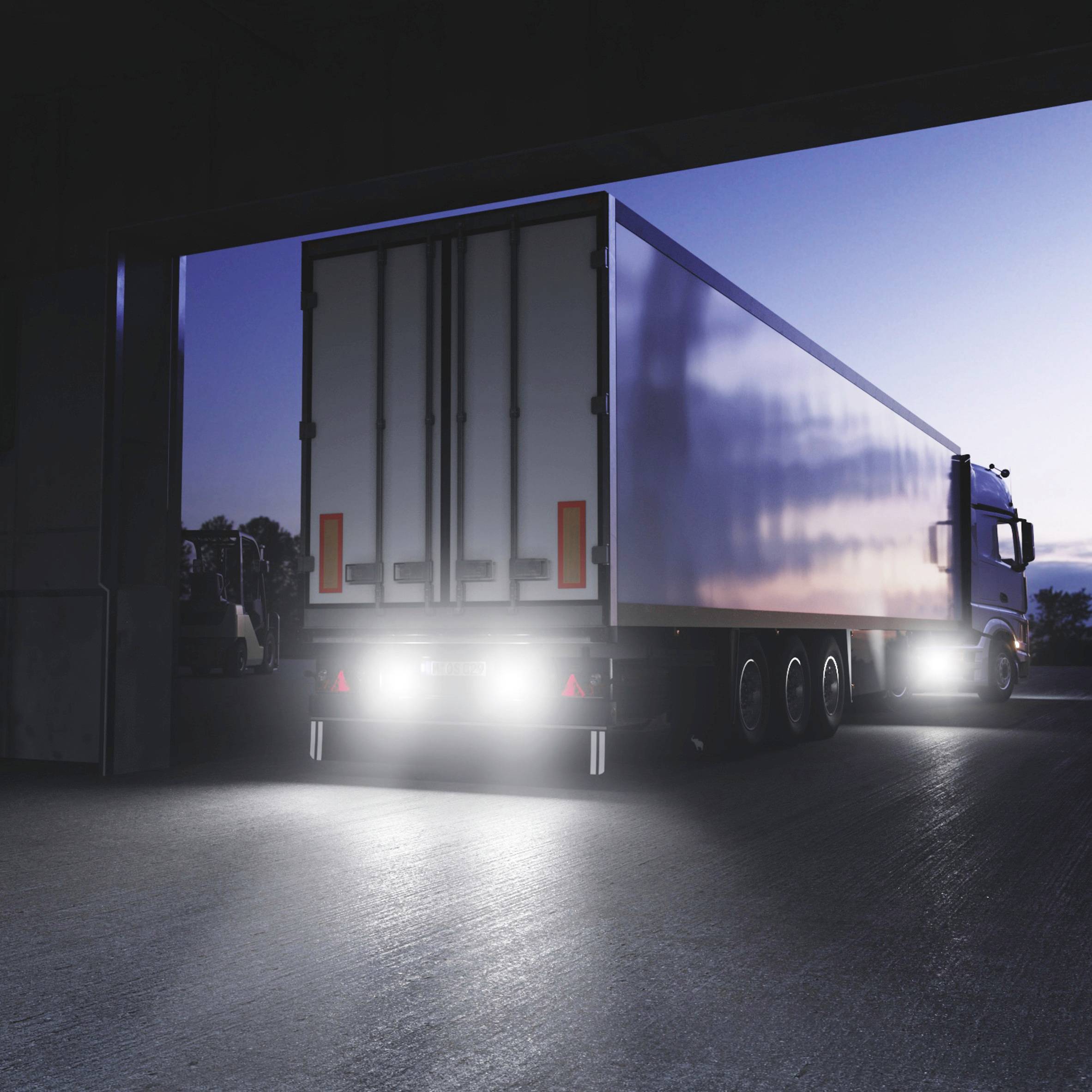 A lorry with an illuminated trailer drives out of a building at dusk.