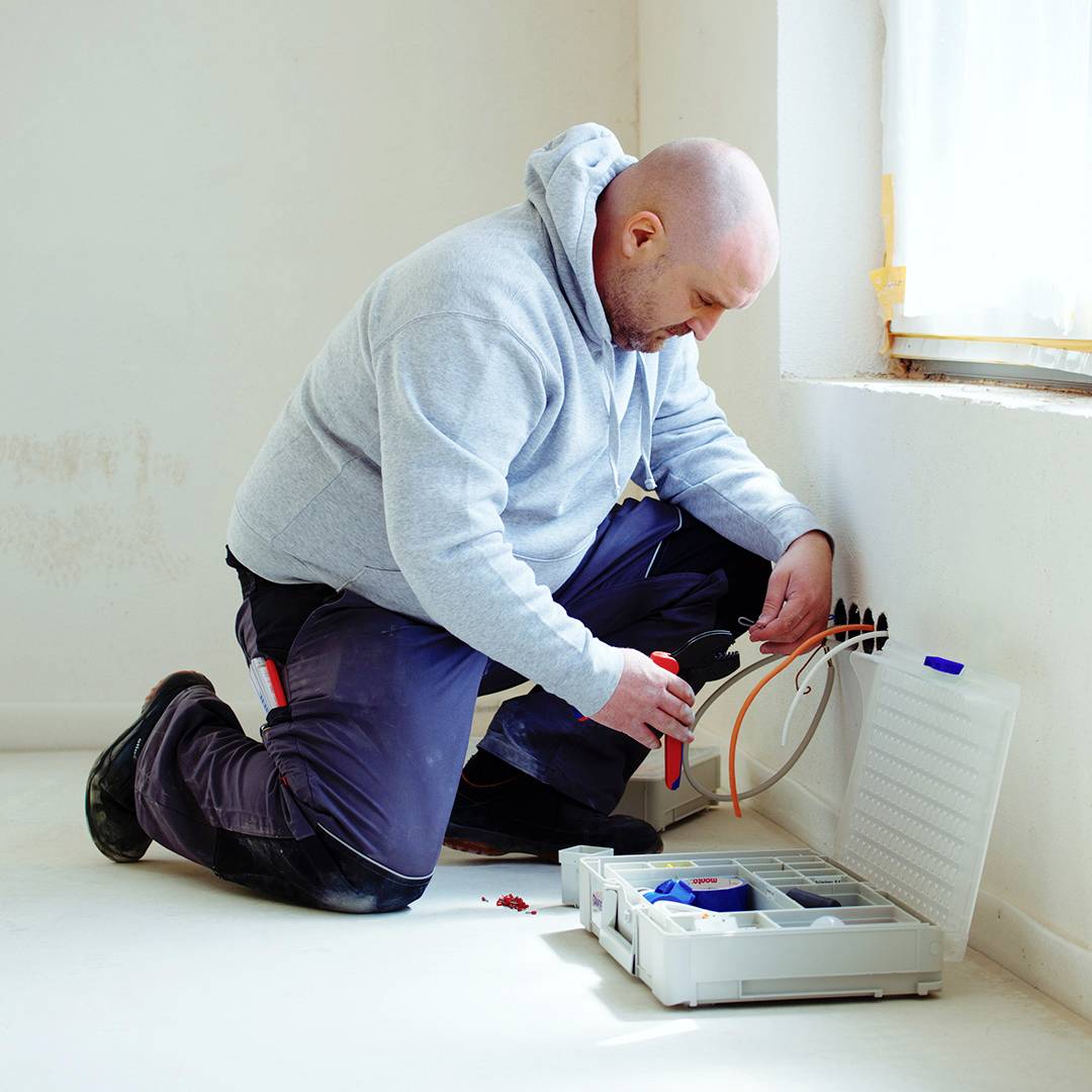 An electrician is kneeling on the floor and working on a cable connection in a flat. Next to him lies an open toolbox.