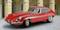 Classic red sports car on a cobbled square in front of a historic building with arches in the background.