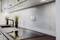 Modern kitchen interior with a dark worktop, black tap, and two potted plants. Bright walls and wall-mounted cabinets.