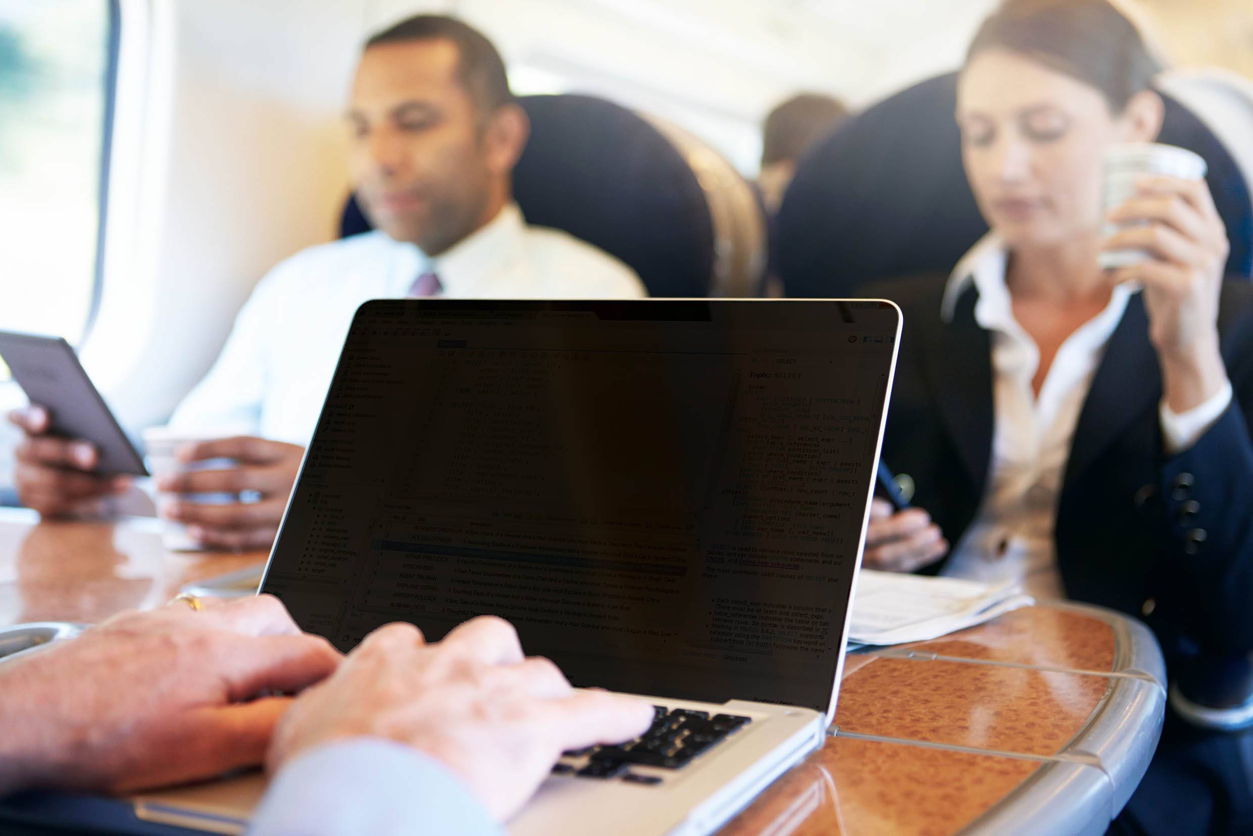People are sitting on the train, working on laptops and tablets. A woman is holding a coffee. A bustling business travel atmosphere.