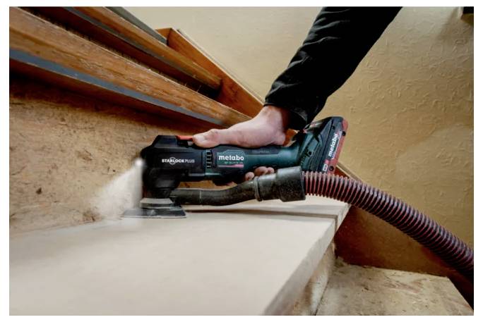 A person uses a hand-held electric sander to smooth the edge of a wooden step, with a vacuum hose for dust extraction.